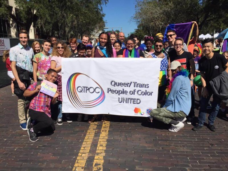 Members of Queer/Trans People of Color United pose around a large banner with the organization's logo.