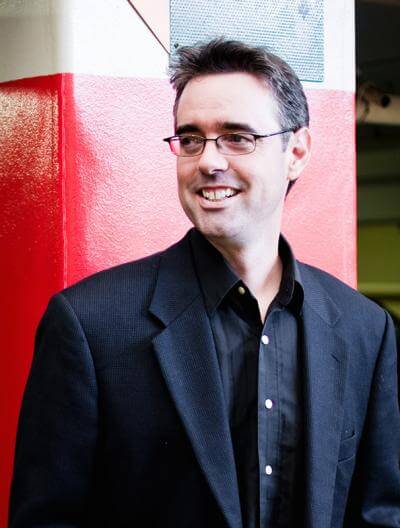 This picture shows Prof. Greg Glancey from Berklee School of Music standing in front of a red pillar in a black suit.