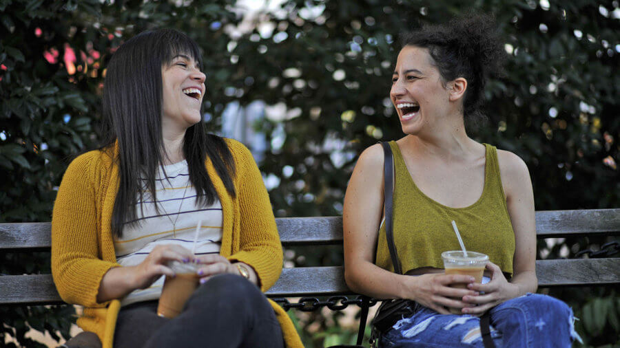 Actresses from Broad City sit on a bench laughing.