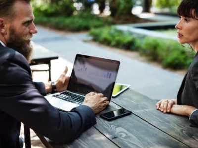 A man and a woman sit across from each other at a picnic table outside. The man has a laptop in front of him and is looking at the woman, who appears to be speaking to him. They are both wearing black blazers and seem to be in an interview.