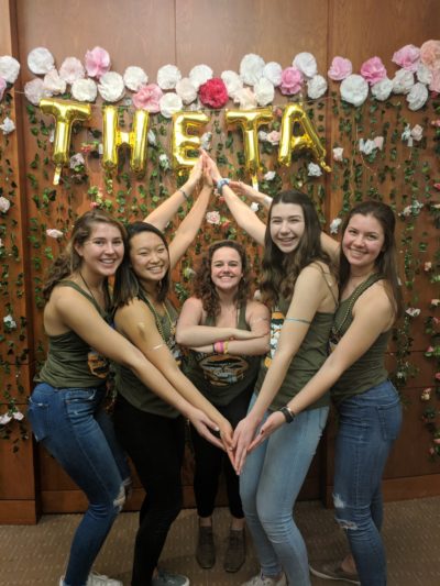 Megan and four other sorority women stand in front of the word Theta with their hands joining to form a kite shape.