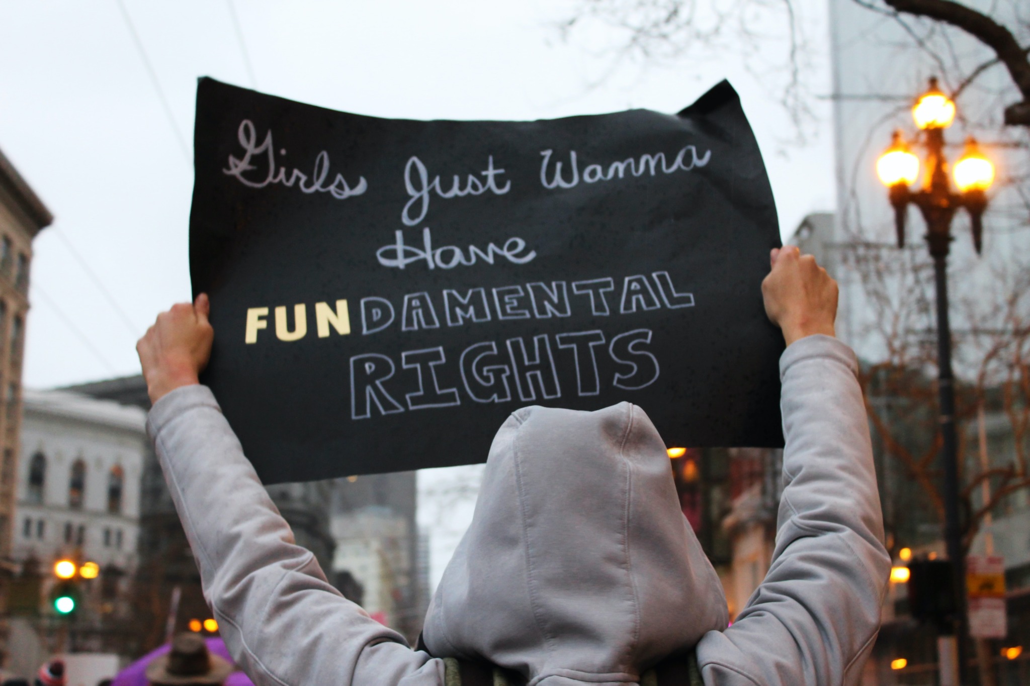  A woman in a grey hoodie holds up a sign which reads 'Girls Just Wanna Have Fundamental Rights' in front of a blurred background of a city street with cars and buildings.