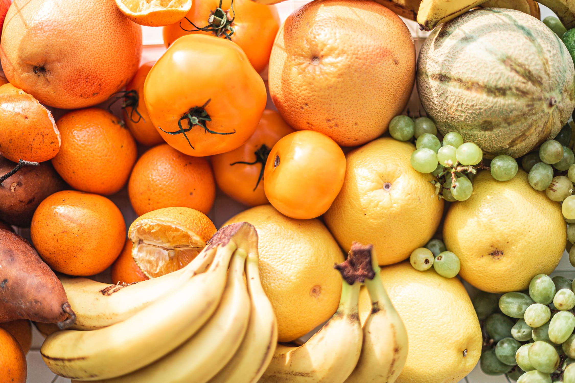 A pile of bananas, grapes, and tomatoes sit in the sun.