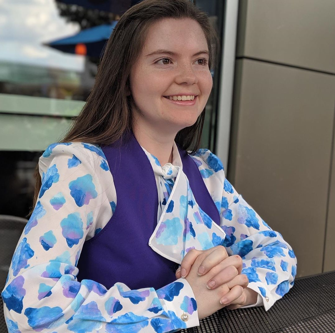 A girl wears a blue shirt and purple vest while folding her hands.