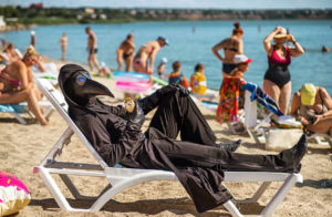 A man in a plague doctor outfit sits on the beach.