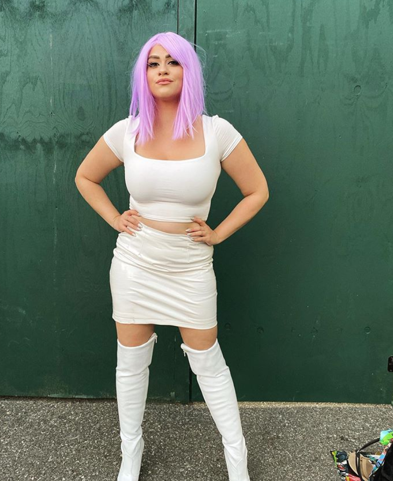 A girl stands in a purple wig and a white outfit.